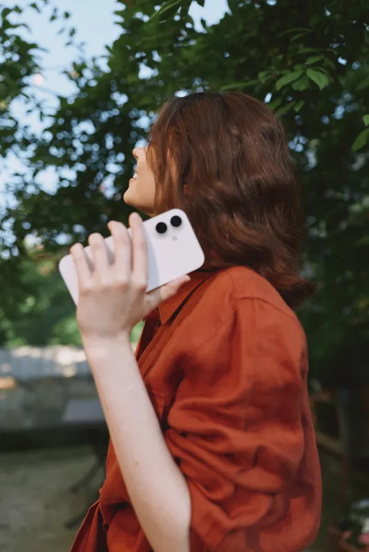 A young woman with long brunette hair talks on a white phone in a sunny outdoor park, wearing an orange blouse and looking to the side with a thoughtful expression