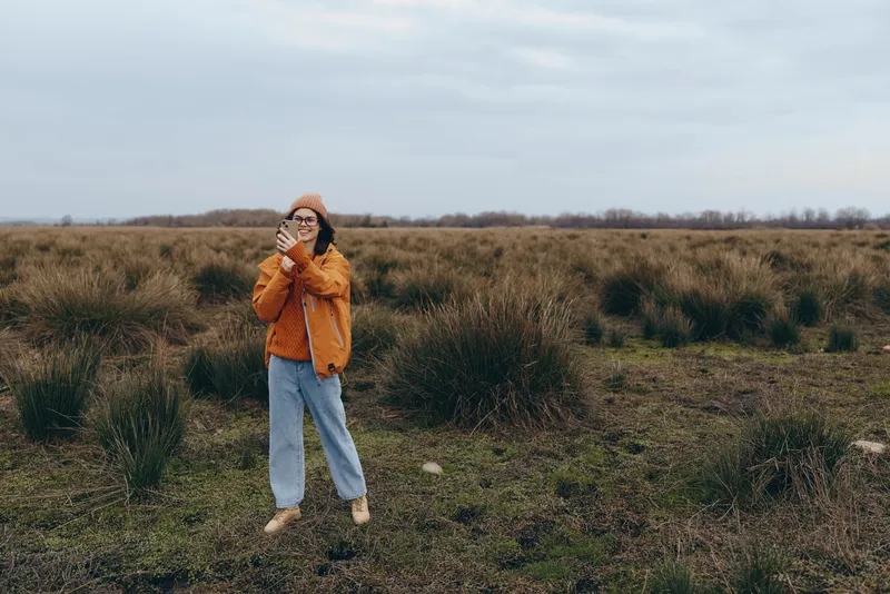 Woman in winter lifestyle stands in a grassy field outdoors, wearing beanie, orange jacket, and scarf, holding the scarf near her face, calm pose under overcast sky, nature landscape, cool wind
