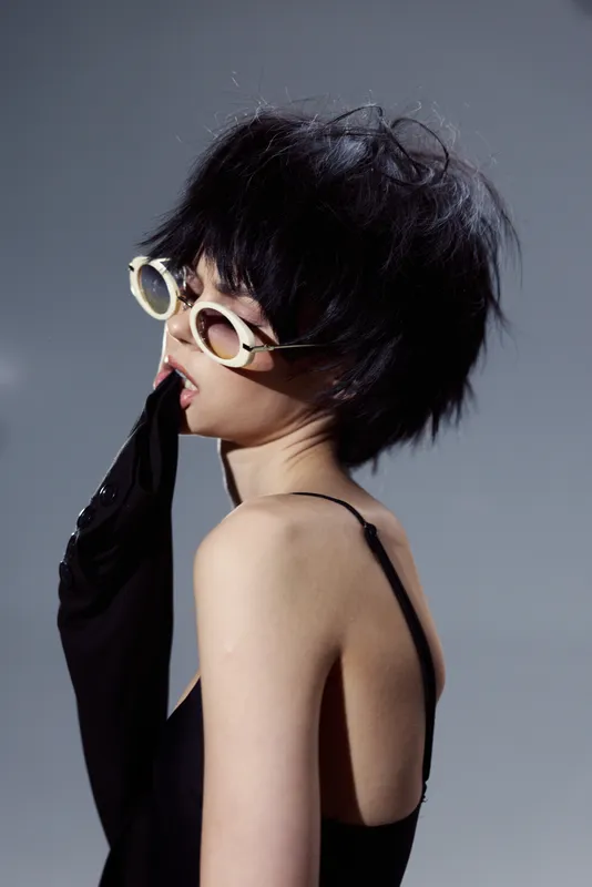 Stylish woman wearing sunglasses and a black dress with a short hairstyle posing against gray background