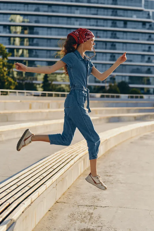 Lifestyle woman jumping in modern boho-western denim street style with a red bandana, casual urban background, film color tones enhancing vibrant energy and motion.