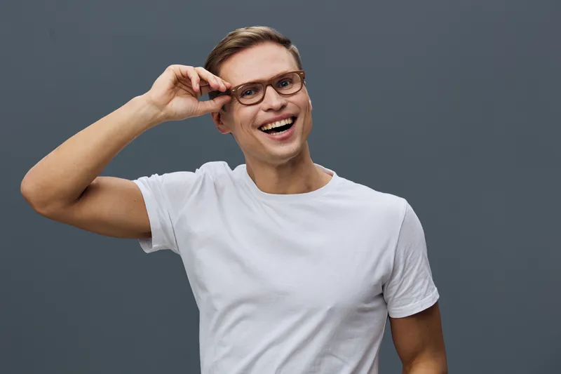 Cheerful man with glasses smiling broadly, casual white t-shirt, holding his glasses with one hand against plain gray background, joyful expression, relaxed pose, modern lifestyle concept.