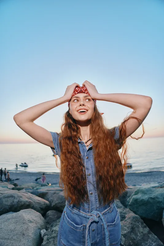 lifestyle woman in modern boho-western denim outfit smiling with long hair and bandana on rocky beach, film color style at sunset with serene ocean background