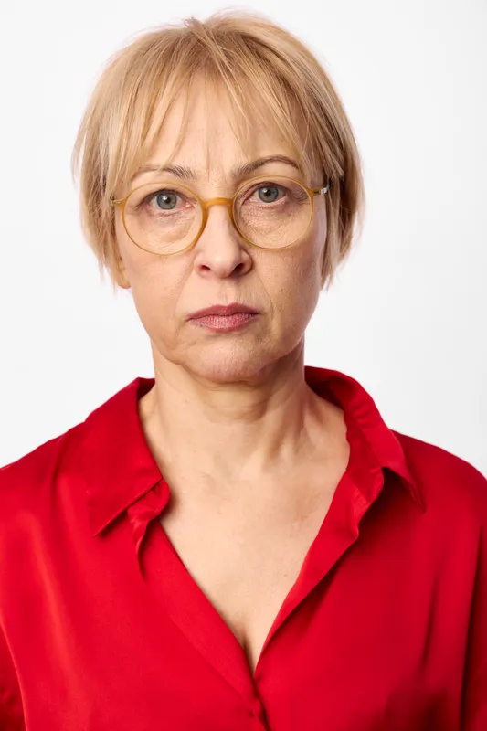 Mature woman portrait with blonde short hair and round glasses, red blouse against white studio background, direct gaze and calm expression, soft lighting, closeup headshot