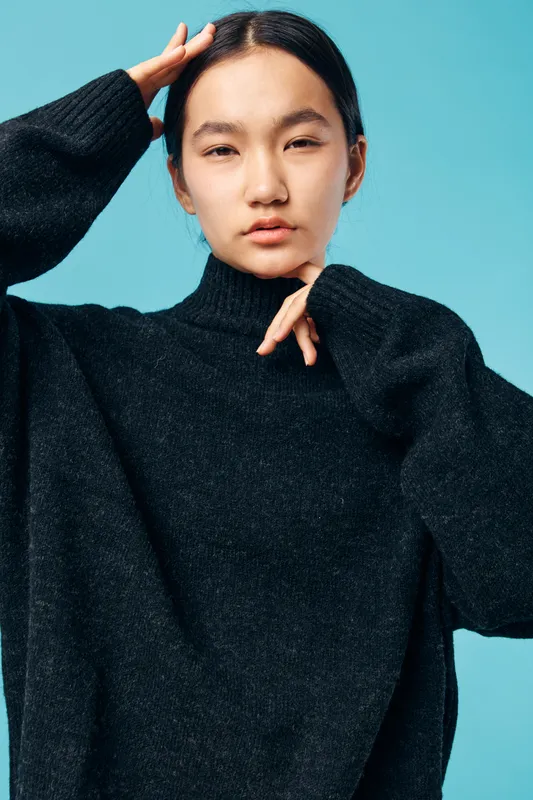 Young woman with black hair wearing cozy dark sweater posing with hand on head against blue background, looking thoughtful and calm with soft natural light.