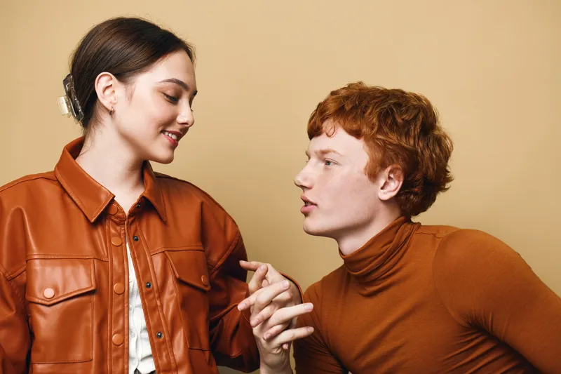 Young man and woman interacting with friendly expressions wearing brown clothing on beige background. Casual communication with focus on faces and hand gestures in conversation.