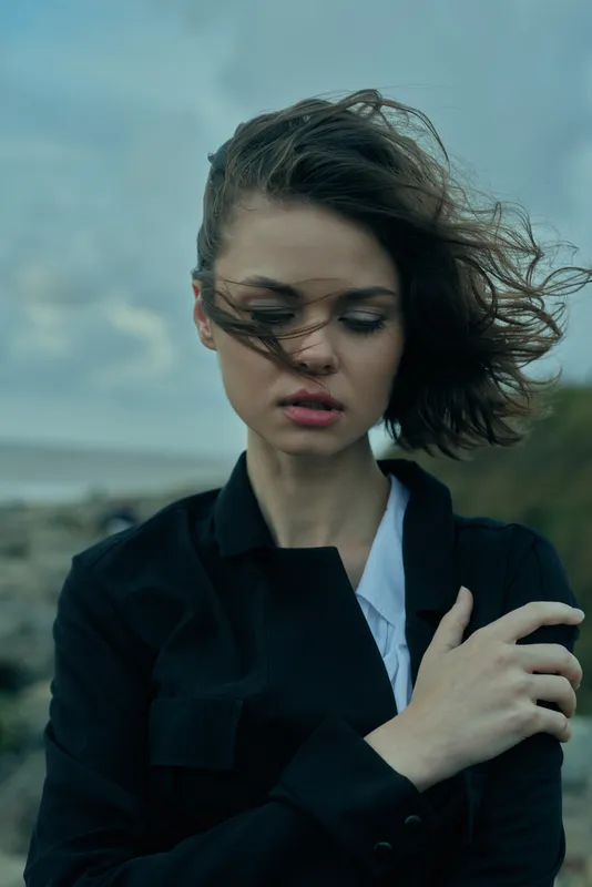 Windy, windswept scene with a young woman in a dark jacket by the coast, hair fluttering, a thoughtful expression, and moody fashion mood in a coastal breeze.