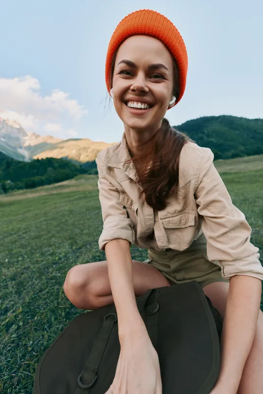 Cheerful woman lifestyle outdoors, smiling on a sunlit mountainside field, casual beige shirt and orange beanie, capturing a carefree moment with nature and wide landscape
