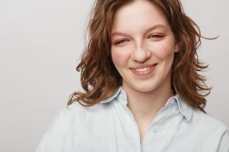 Smiling young woman with wavy brown hair wearing a white shirt looks down with a gentle expression on a plain light background. Concept of natural beauty and casual lifestyle.