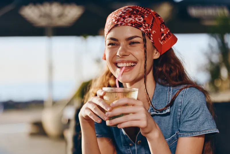 Lifestyle woman with modern boho-western denim street style enjoying a drink outdoors, smiling brightly in warm film color tones, capturing relaxed youthful energy.