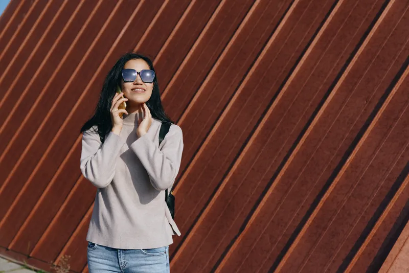 Woman in casual sweater and jeans talks on a mobile phone against a striped wooden wall, sunglasses, outdoor urban setting, lifestyle image focused on communication and fashion.