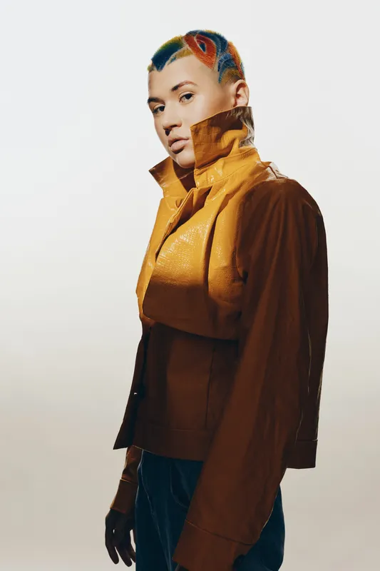 Stylish woman with colorful short hair wearing a brown leather jacket and gloves, posing confidently against a plain background in a contemporary fashion portrait.