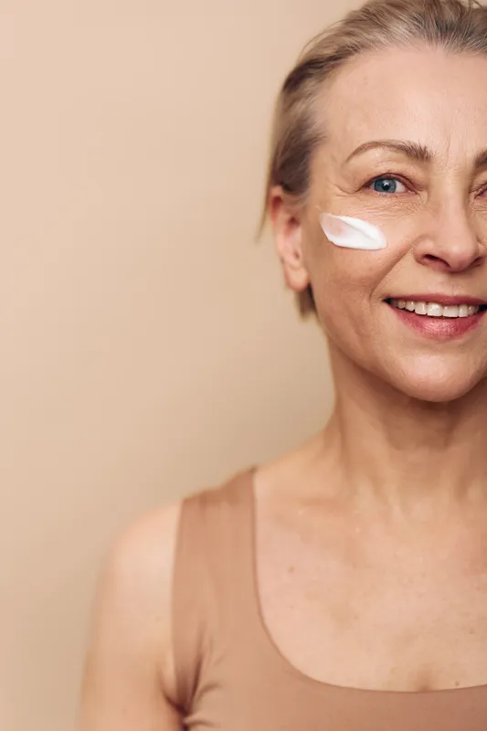 Smiling mature woman with short hair applies white face cream on cheek. Close-up on warm beige background. Concept of skincare, beauty routine, self-care, and natural aging with healthy skin.