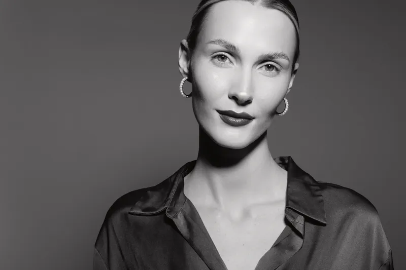 Portrait of a young white woman with pale skin and light hair wearing hoop earrings and a dark blouse. Studio shot in black and white with soft lighting and neutral expression.
