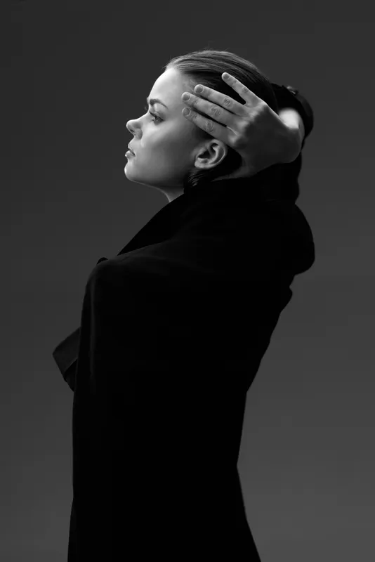 Black and white portrait of a thoughtful woman with hands in hair wearing a dark coat in studio lighting.