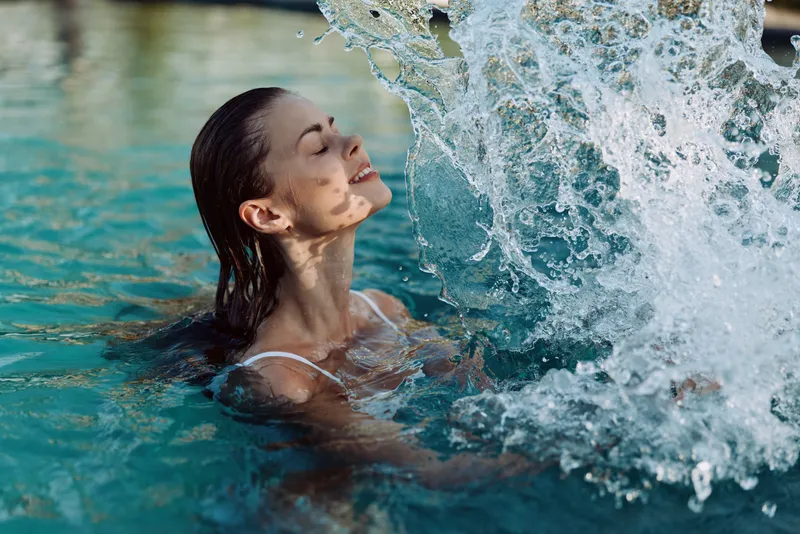 Cheerful woman enjoying a splash in a clear pool, serene smile, sunlit water droplets, relaxed posture, outdoor setting, vibrant blues, natural light, carefree moment, lifestyle scene, refreshing