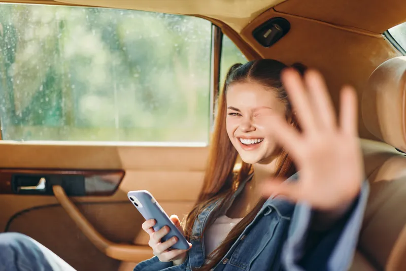 Cheerful young woman enjoying her time in a car, smiling and waving while using her smartphone, showcasing carefree emotions and a fun day out.