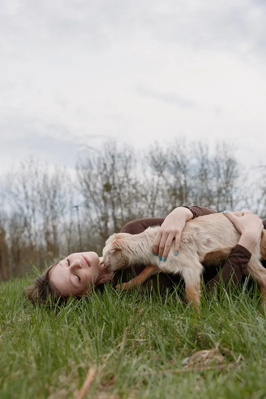 A girl lies on the grass outdoors, embracing a curious goat kid. The scene conveys affection, relaxation, and a natural bond in a countryside setting with light and calm atmosphere.