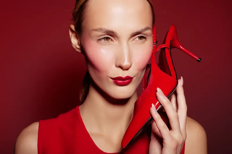 Woman in red dress holds red high heel shoe near face on red background. Fashion concept with bold makeup and stylish accessories in studio lighting.
