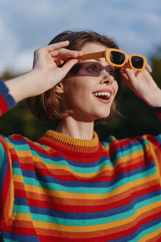 Woman in sunglasses and a rainbow sweater shows a bright smile in a portrait outdoors, joyful fashion moment with natural light, colorful styling and candid expression.