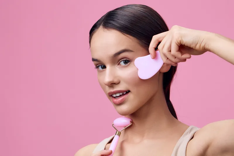 Young woman using pink skincare tools on her face against a pink background, showcasing a vibrant, youthful skincare routine