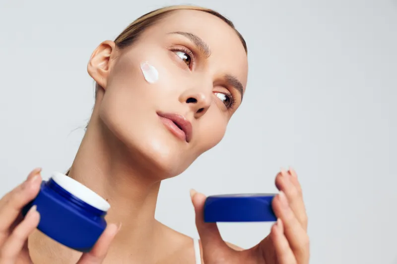 Young woman applies face cream on cheek holding blue jar with lid in hand looking away with smooth skin and natural makeup isolated on gray background in skincare concept.