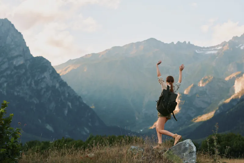 Young woman with a backpack joyfully raises her arms while standing on a rock in a mountainous landscape during daylight.