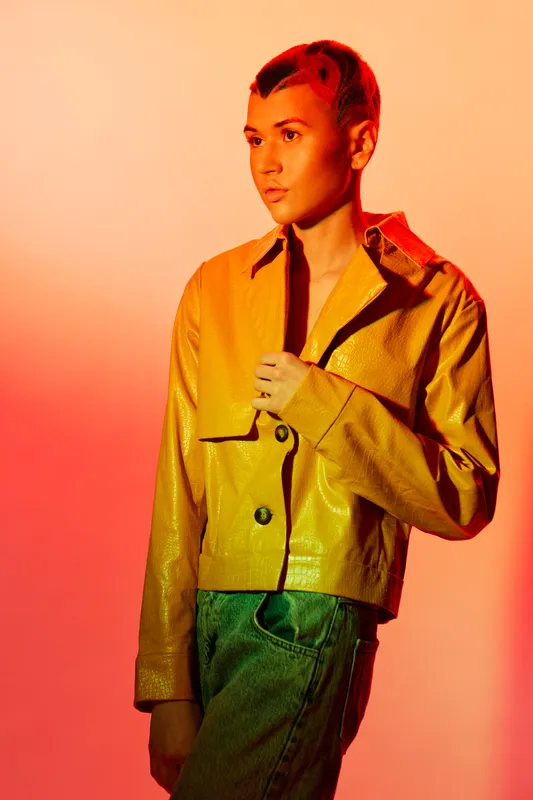 young woman with short hair wearing yellow jacket and jeans standing against orange background, expressing thoughtful mood in stylish casual outfit