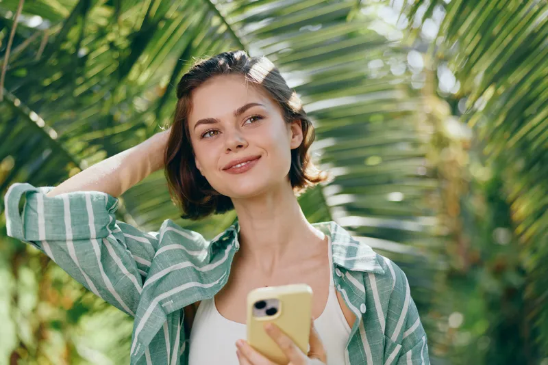 Young woman outdoors, smiling softly while holding a smartphone. Casual striped green shirt with a leafy, sunlit backdrop.
