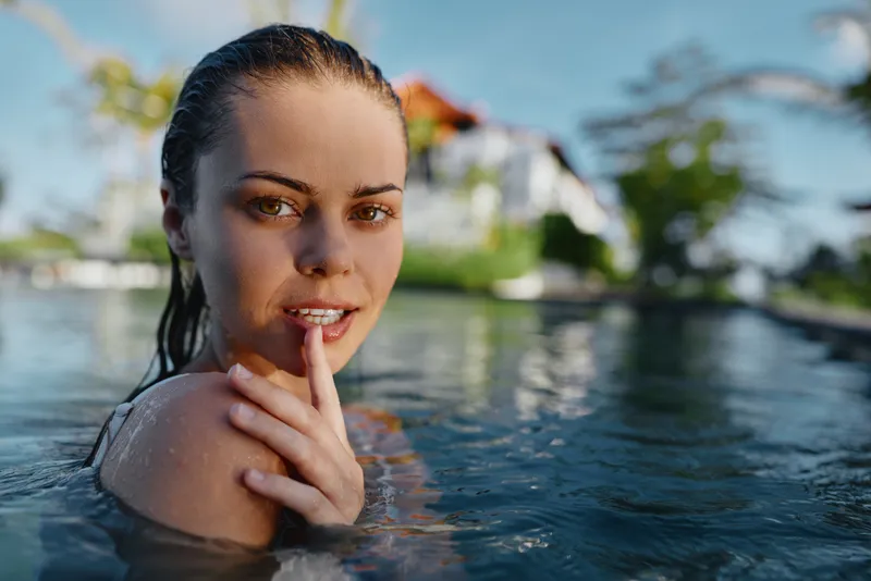 Young woman with wet brunette hair relaxes in a swimming pool on a sunny day, looking thoughtfully at the camera with her hand near her face, creating a calm and natural mood.