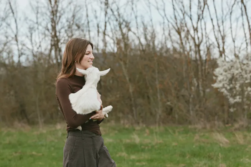 Joyful woman outdoors, smiling while holding a white goat kid in a sunlit field, casual outfit, gentle breeze, natural colors, candid lifestyle moment