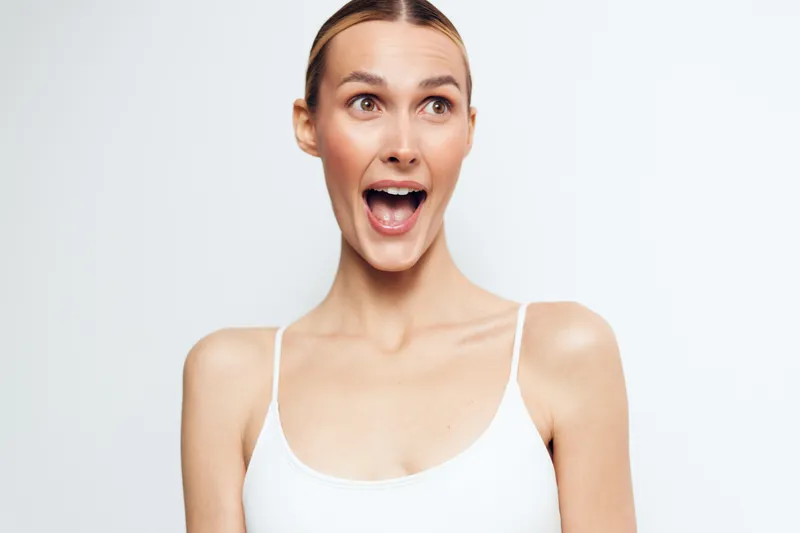Young woman with surprised expression wearing a white tank top stands in a bright studio against a clean background, capturing a candid, upbeat moment with natural light and a fresh composition
