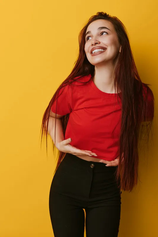 Young woman with long hair wearing red shirt and black pants smiling and looking up on yellow background. Happy female model posing with hand gesture in casual outfit for lifestyle portrait.
