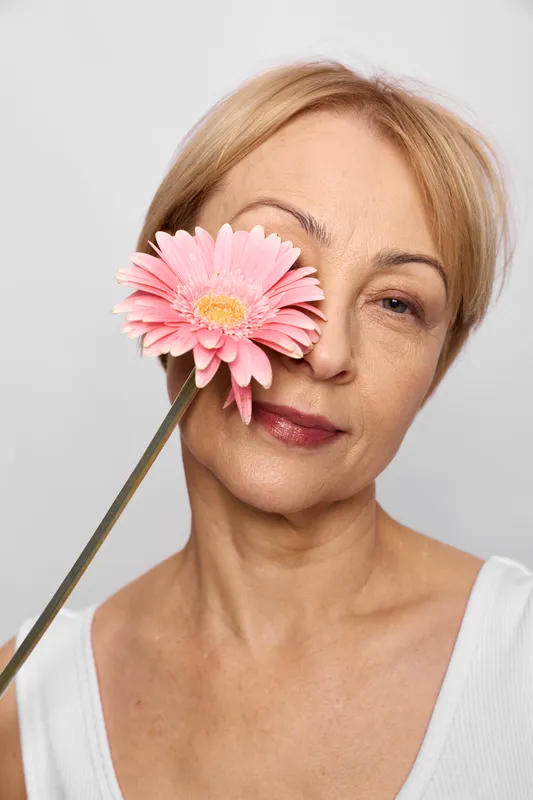 Blonde woman portrait in studio, smiling softly, holding a pink gerbera flower near her eye, closeup headshot on white background, calm expression, clean light, minimal beauty