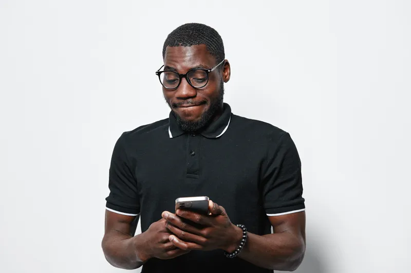 African American man with dark skin wearing glasses and black polo shirt using smartphone for texting or browsing on white background. Casual adult male focused on mobile device.