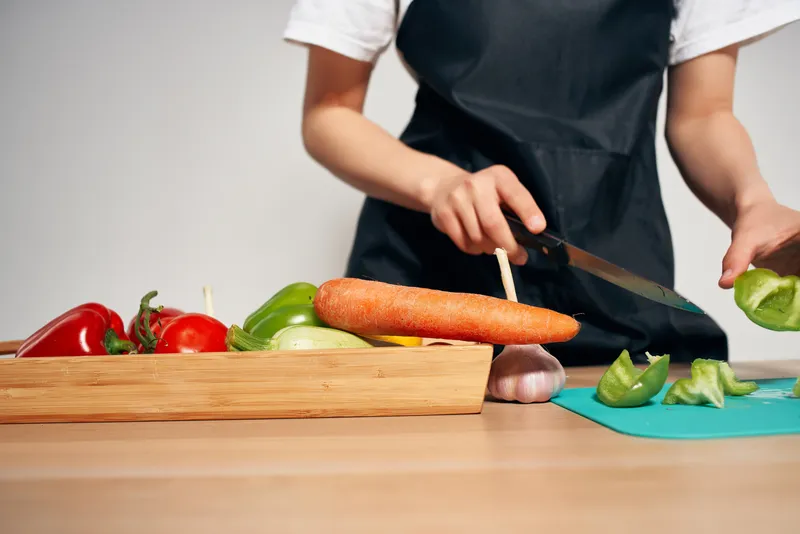 Женщина в черном фартуке нарезает овощи на кухне, домохозяйка готовит еду