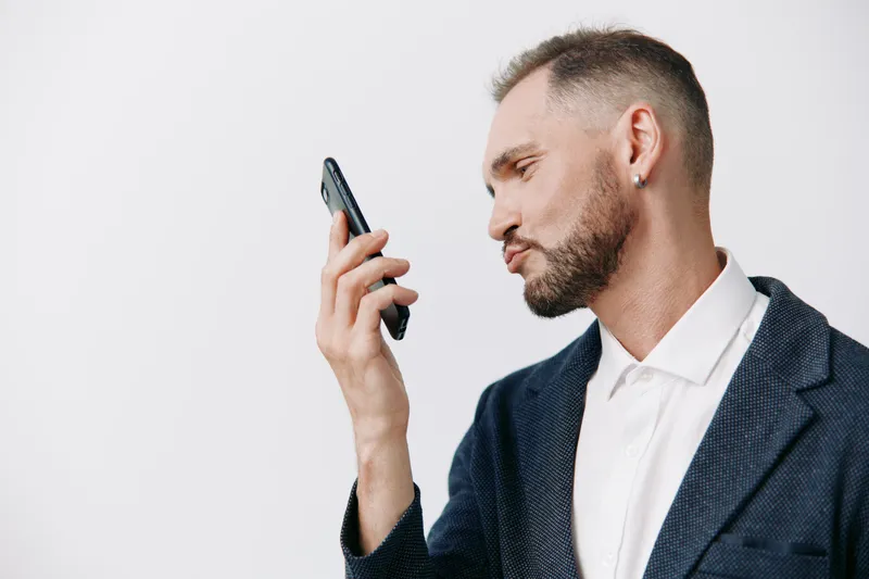 A professional man in a business suit examines a smartphone with focused emotion against a clean isolated colored background. The calm, confident pose conveys concentration, communication, and