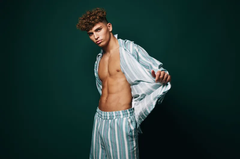 Young man with curly hair wearing striped pajamas posing against dark background, showing toned torso and casual style in studio lighting for fashion or lifestyle.