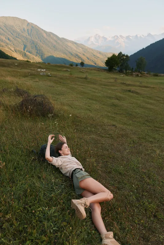 Cheerful woman outdoors lying on grass, sunlit valley scene, relaxed pose, casual outfit, distant mountains, warm breeze, natural colors, serene lifestyle moment