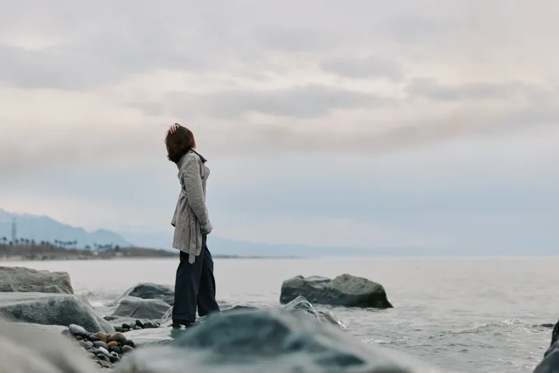 A woman stands on rocky shore facing calm sea under cloudy sky. She wears a long cardigan and looks contemplative in cool outdoor landscape. Concept of solitude, nature, and reflection.
