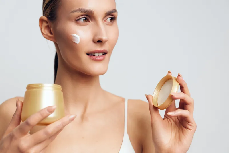 A young woman in a white tank top applies cream on her cheek holding an open jar of skincare product, demonstrating a daily beauty routine for healthy skin and hydration.