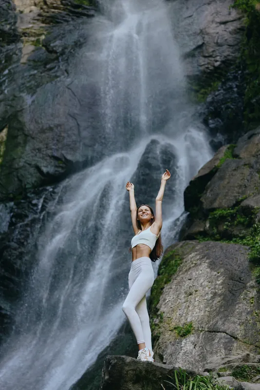 woman standing near waterfall with arms raised wearing white sportswear, surrounded by rocky nature and flowing water in an outdoor setting.