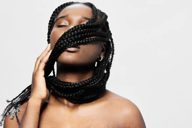 Woman with braided hair, softly embracing her hairstyle against a neutral background, showcasing beauty and confidence in a serene, artistic portrait