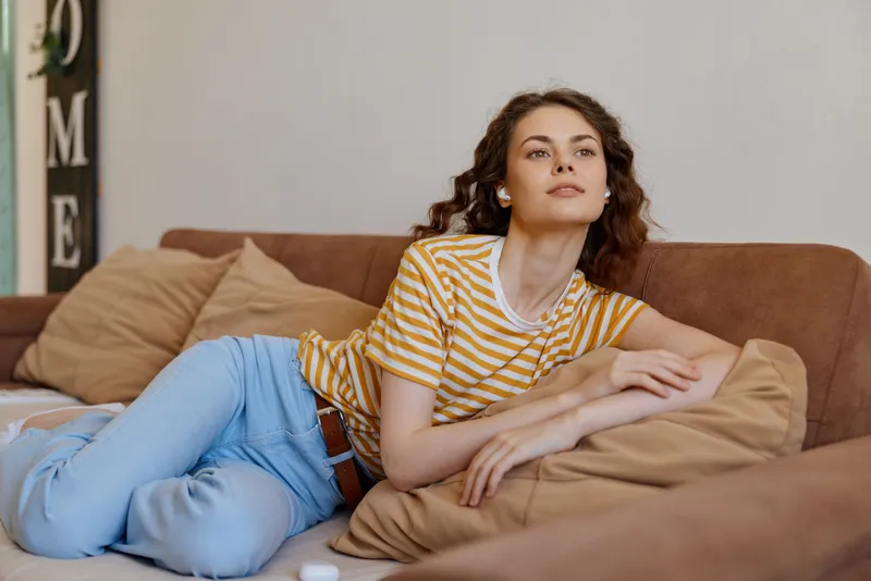 A relaxed woman lounges on a beige sofa at home, wearing a striped tee and jeans, soft light filling a cozy living room, conveying casual lifestyle and comfort.