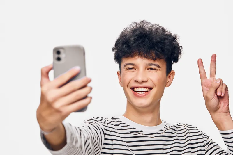 young man taking selfie smiling with peace sign wearing striped shirt on white background, happy expression, casual style, modern youth culture