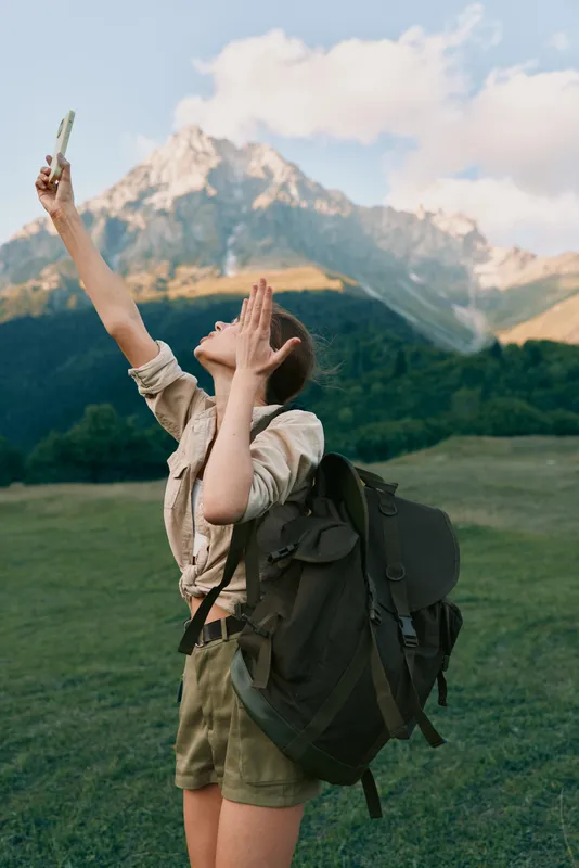 One woman hiker with a backpack cheers toward the sky in a mountain meadow, capturing the trail view with a phone, exuding excitement and freedom.