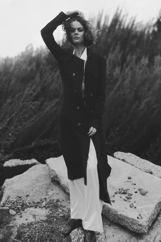 Woman in a long black coat over a white dress stands on rocky terrain in a moody monochrome outdoor shot, windswept hair, minimal contrast and strong silhouette for a fashion editorial.