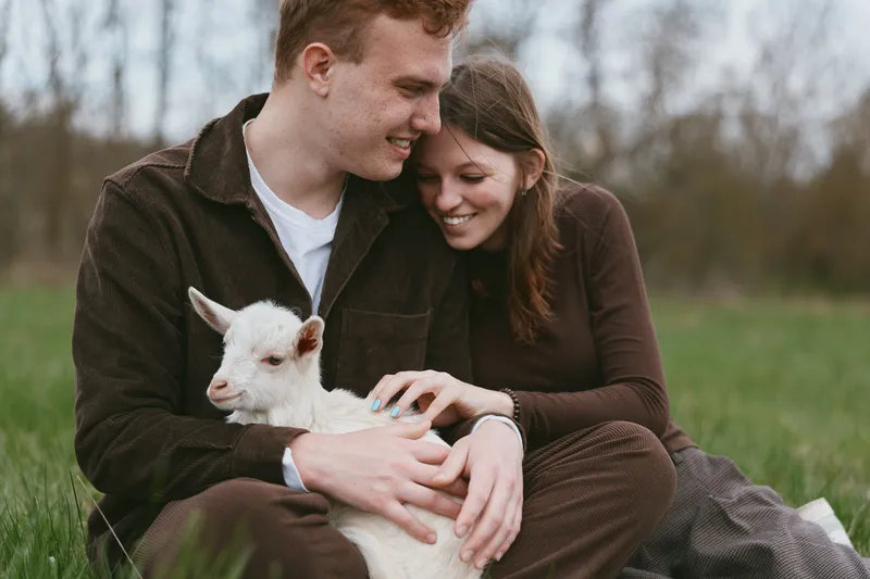 Young couple sitting on grass gently holding a white baby goat in their arms during a cozy outdoor moment in early spring with soft natural light and casual clothing.