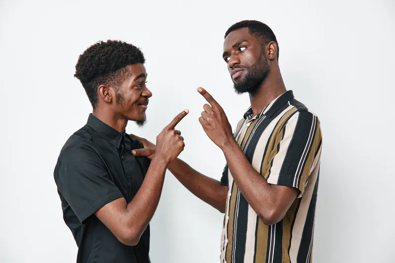 Two young black men with dark skin standing and pointing fingers at each other in friendly interaction on white background. Casual clothing and positive facial expressions.