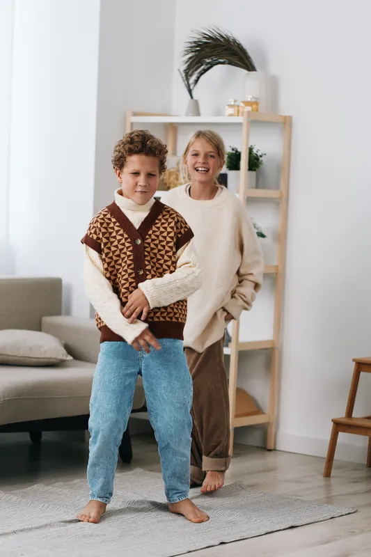 Two smiling children stand indoors on a light rug, posing playfully in cozy knit sweaters and jeans, capturing a warm, casual family moment.