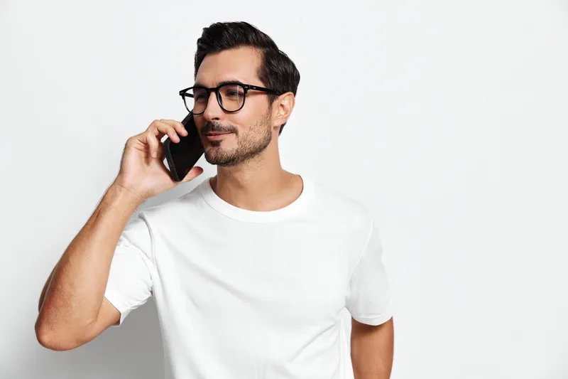 Smiling man talking on mobile phone wearing reading glasses, casual white t-shirt, isolated on plain solid background. Lifestyle concept in modern communication.